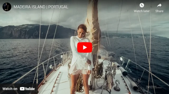 A video thumbnail of a woman on a sailboat, dressed in white, standing at the bow and looking at the horizon. The scene conveys freedom and luxury while exploring Madeira's coastline.